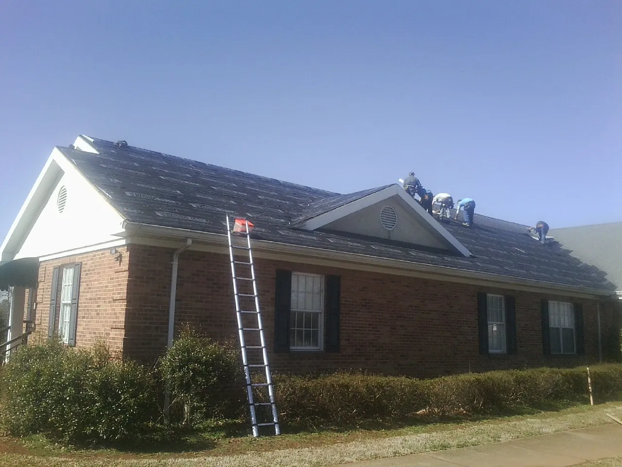 Crew tearing shingles off a brick ranch roof with a ladder set — Emergency Roof Repair work by our crew in Tintah