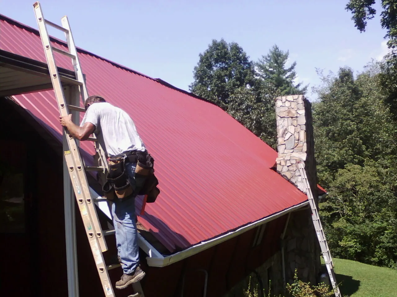 Roofer on a ladder beside a red metal roof and stone chimney — Soffit & Fascia Repair by Roofing Brothers in Tintah